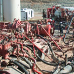 Industrial oilfield site with red machinery and extensive piping; workers in hard hats inspecting equipment.